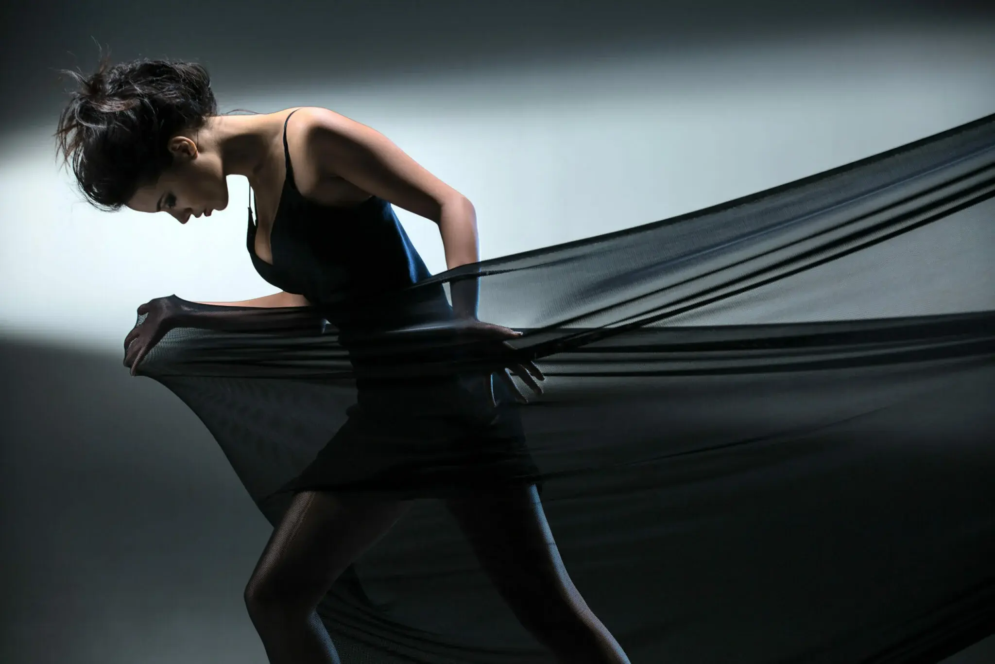 Graceful brunette stretches stretch mesh fabric in front of you on the background of bright ray of light in the studio. Fashion photography. The girl in a dark dress short.